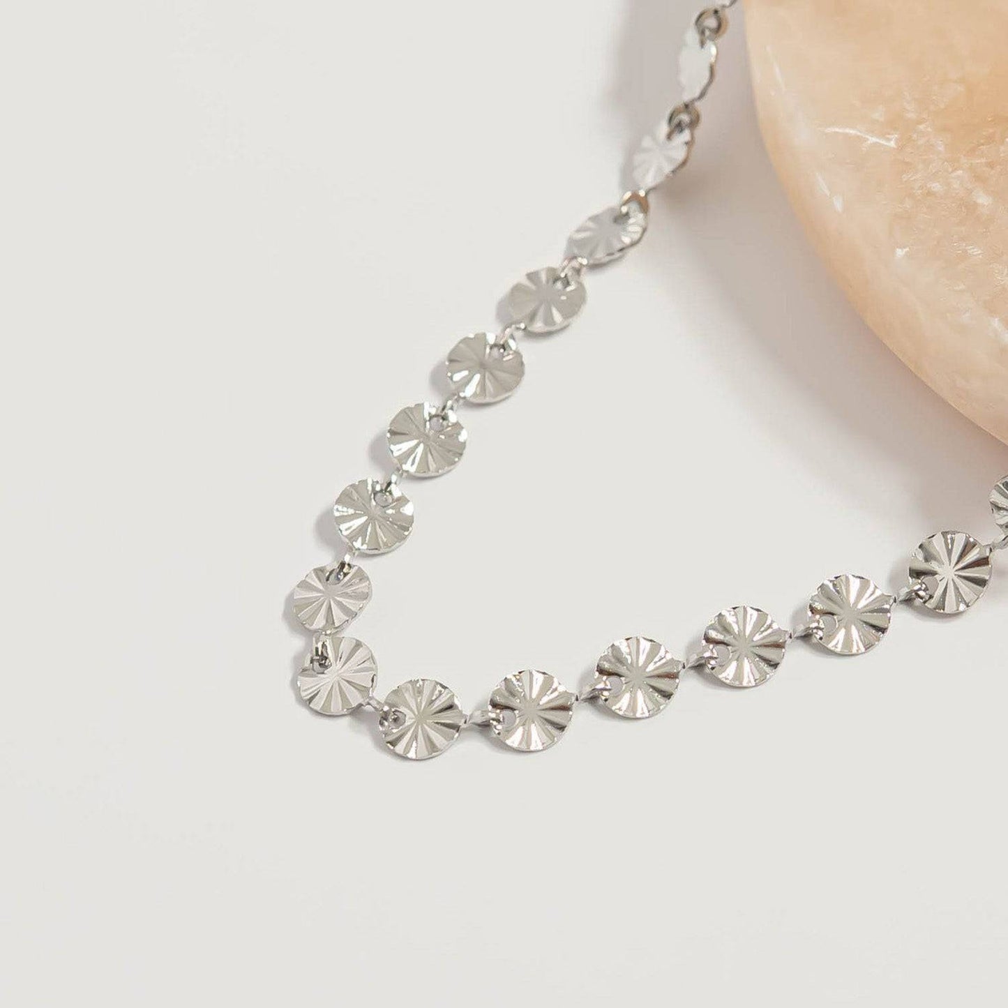 Close-up of silver sunburst disc chain necklace with reflective hypoallergenic waterproof finish displayed on neutral stone