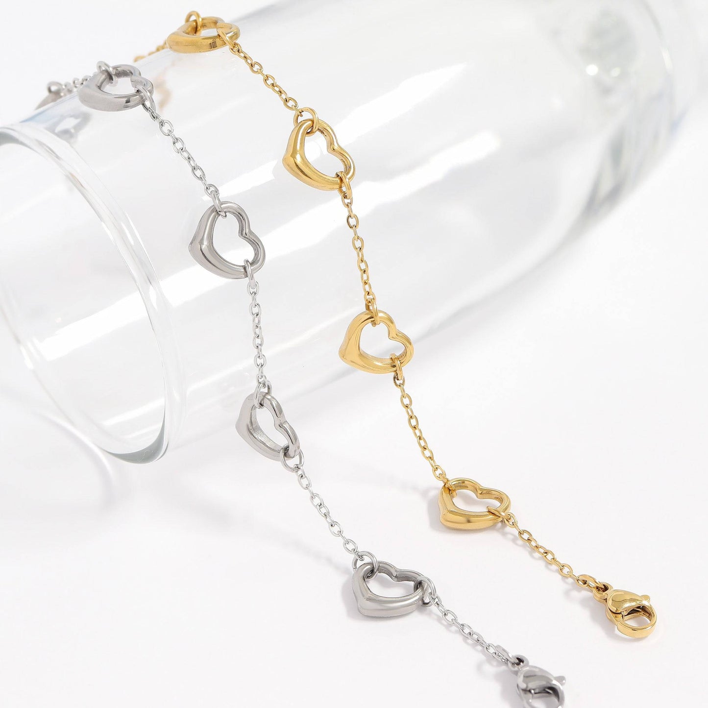 Dainty gold and silver heart bracelets displayed on a glass, featuring open-heart charms and waterproof, tarnish-free stainless steel chains.