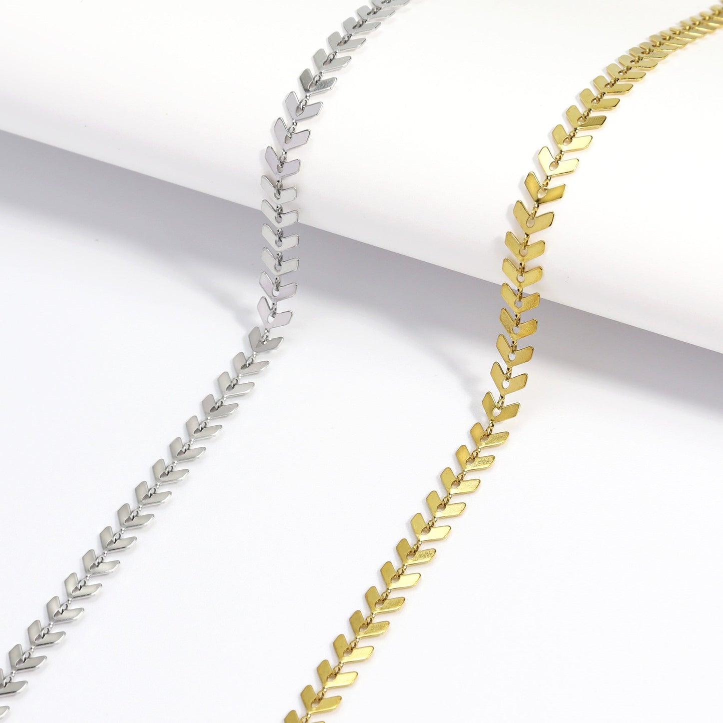 Flat lay display of gold and silver herringbone chain necklaces showcasing the sleek, reflective finish and elegant chevron design against a minimal white background.