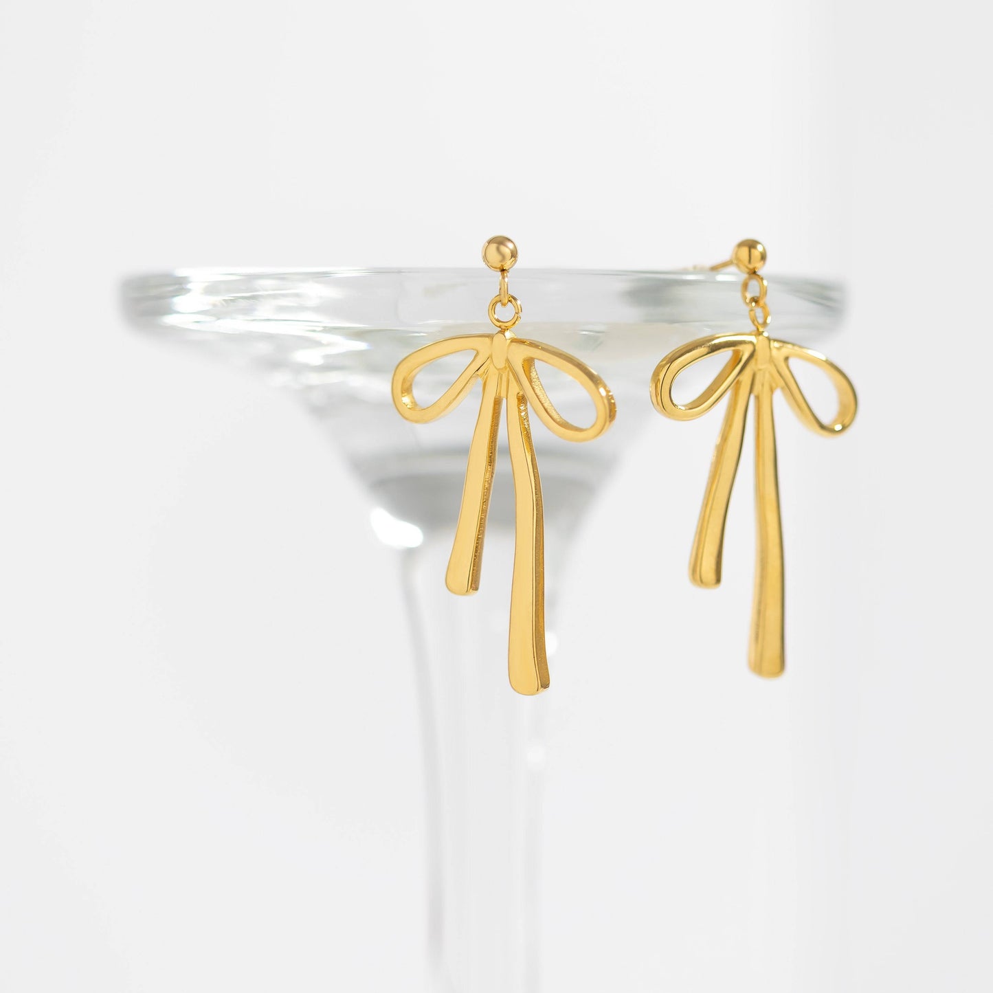 Pair of gold bow drop earrings styled on a clear glass rim. Elegant ribbon design, hypoallergenic and waterproof—perfect for everyday luxury by Lauren Riley Jewelry.