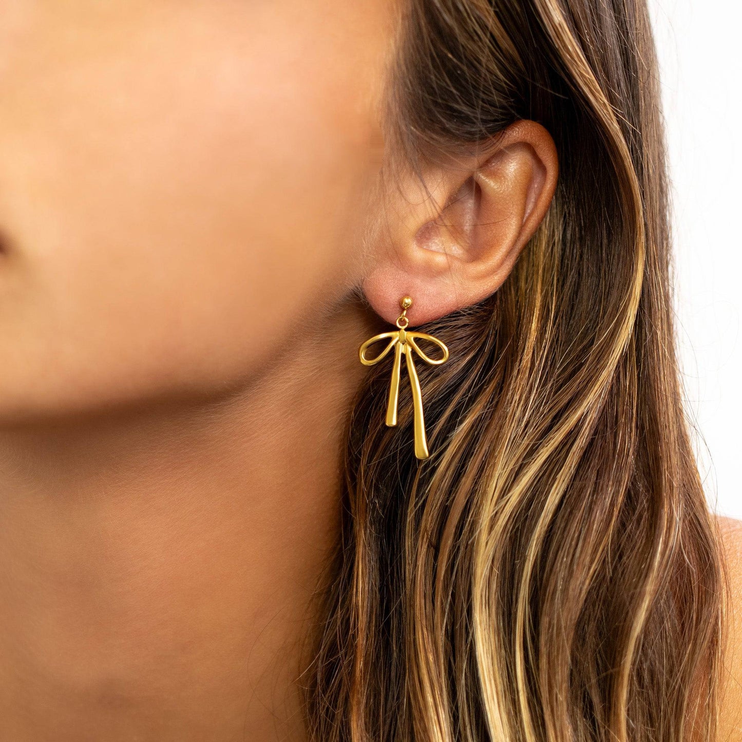 Close-up side view of a woman wearing gold bow drop earrings with a ribbon-inspired design. Hypoallergenic and waterproof earrings by Lauren Riley Jewelry, styled for everyday wear and elegance.