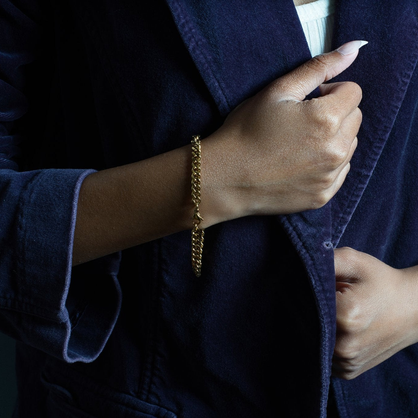 Close-up of a woman's wrist adorned with a gold Cuban chain bracelet, styled with a deep purple velvet blazer for an elegant and bold look.