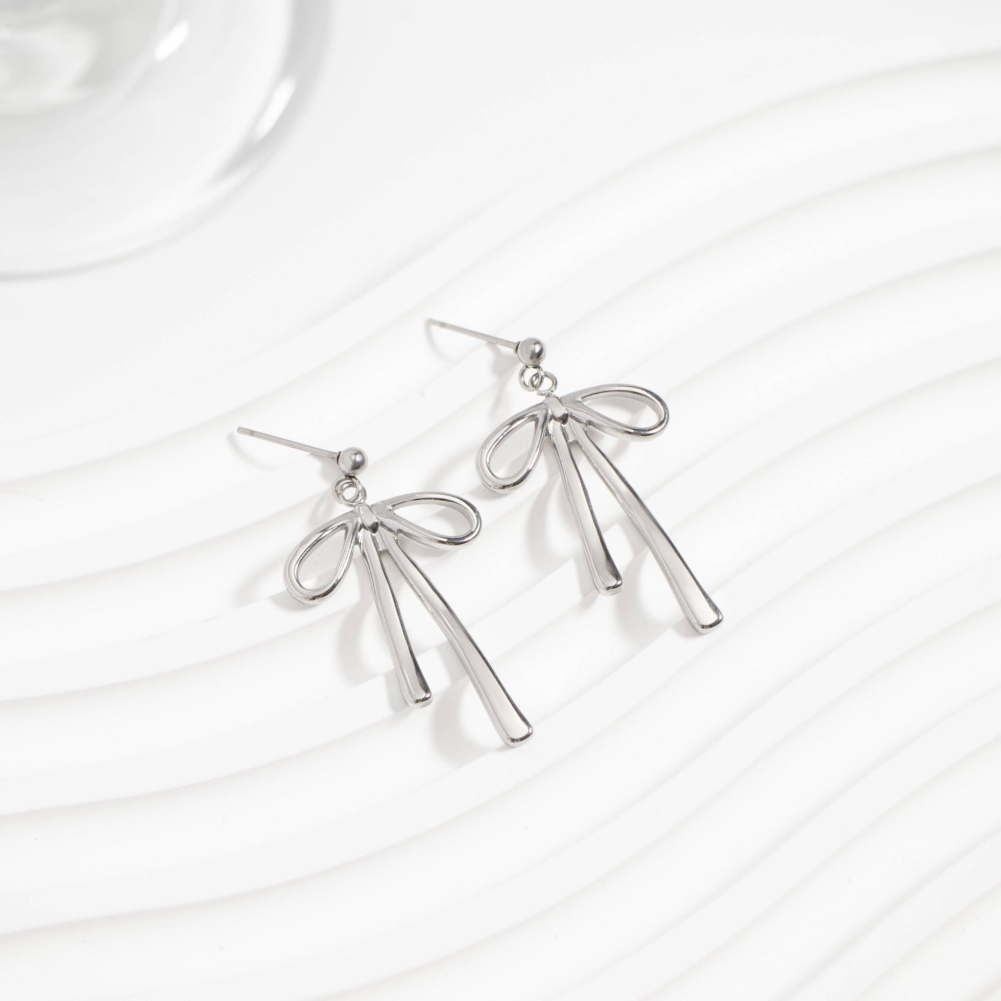 Pair of silver bow earrings displayed on a wavy white background. Dainty ribbon-inspired design that's hypoallergenic, tarnish-resistant, and perfect for everyday elegance from Lauren Riley Jewelry.