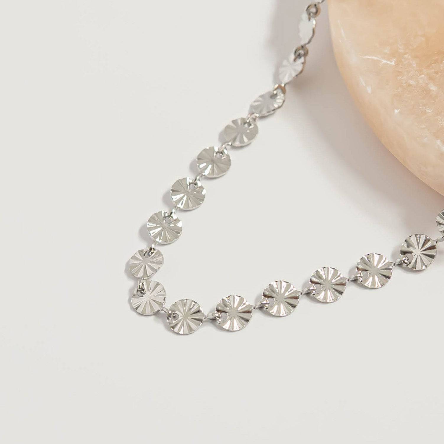 Close-up of silver sunburst disc chain necklace with reflective hypoallergenic waterproof finish displayed on neutral stone