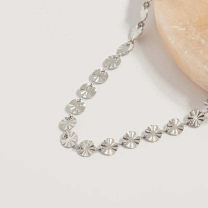Close-up of silver sunburst disc chain necklace with reflective hypoallergenic waterproof finish displayed on neutral stone