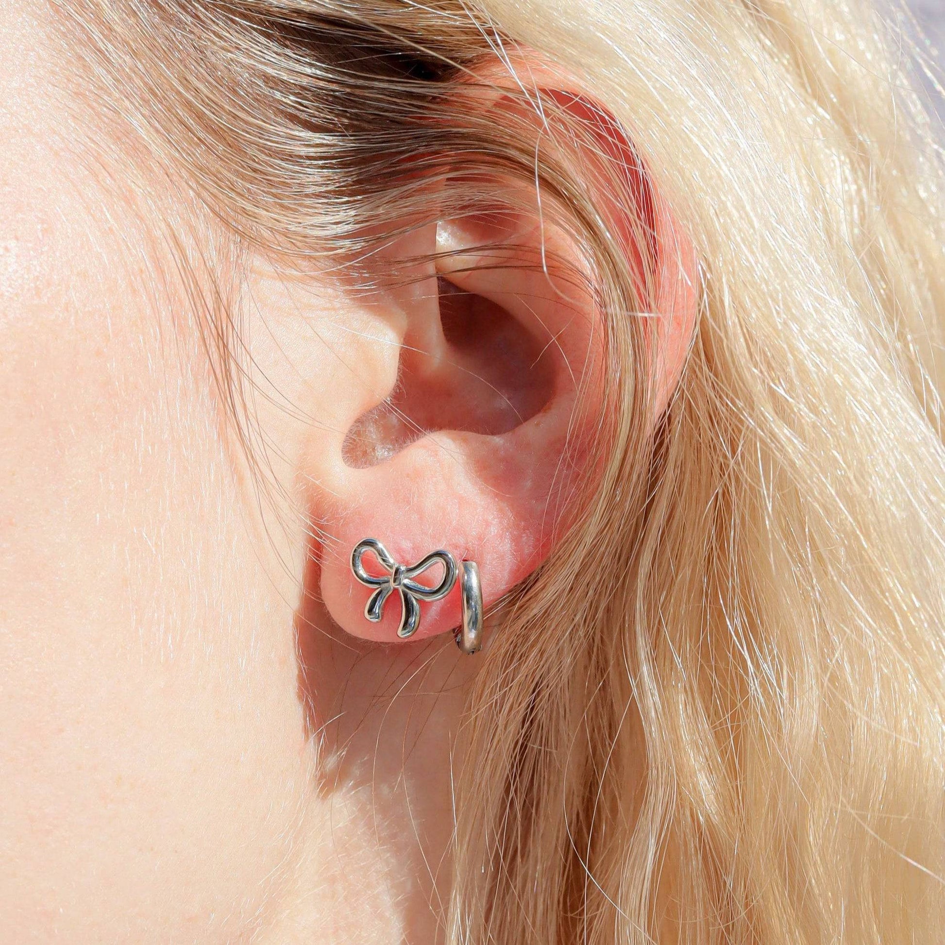Close-up of dainty silver bow stud earrings worn on ear, minimalist hypoallergenic jewelry for everyday wear