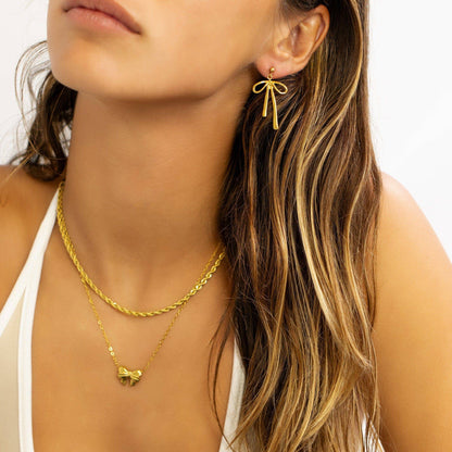 Woman wearing gold bow drop earrings and a layered gold necklace featuring a puff heart charm. Chic, waterproof, and hypoallergenic pieces styled by Lauren Riley Jewelry for a timeless everyday look.