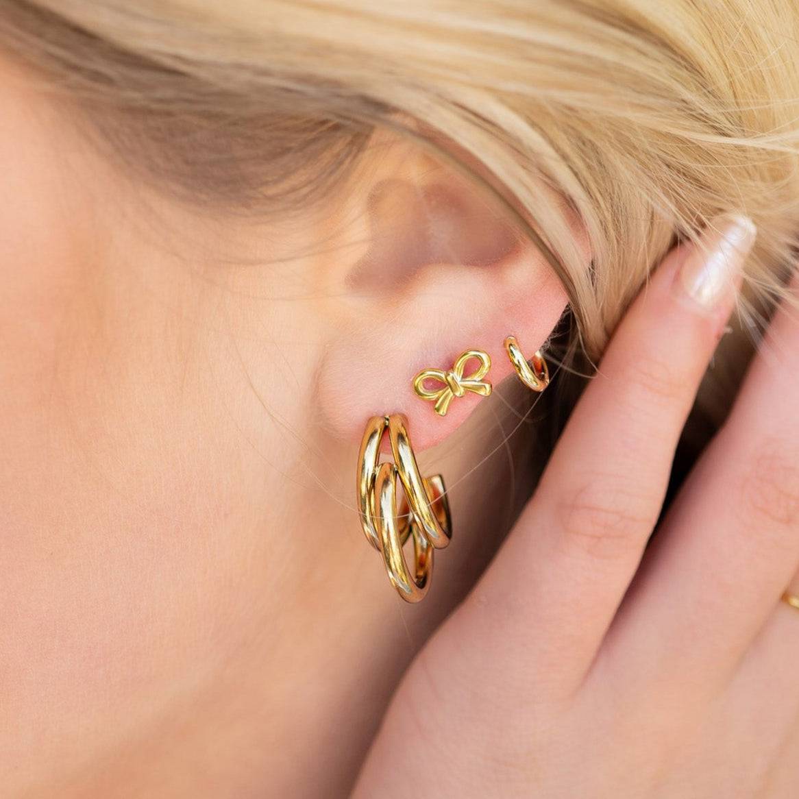 Dainty gold bow stud earrings paired with chunky gold hoops in a trendy stacked ear styling on blonde woman