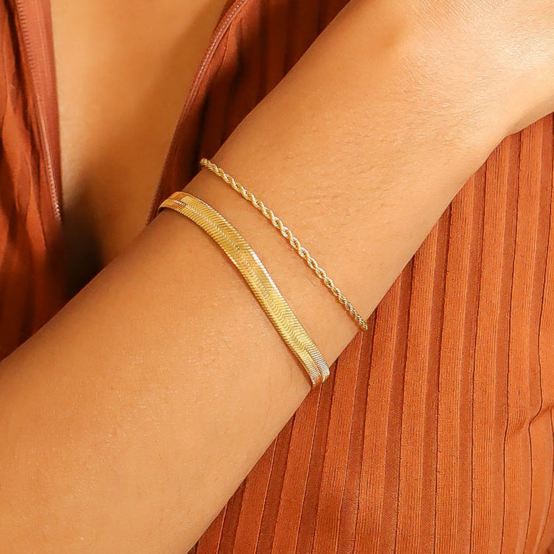 Layered look featuring a silver rope chain bracelet stacked with a bold textured cuff, worn on wrist with a rust-orange ribbed top