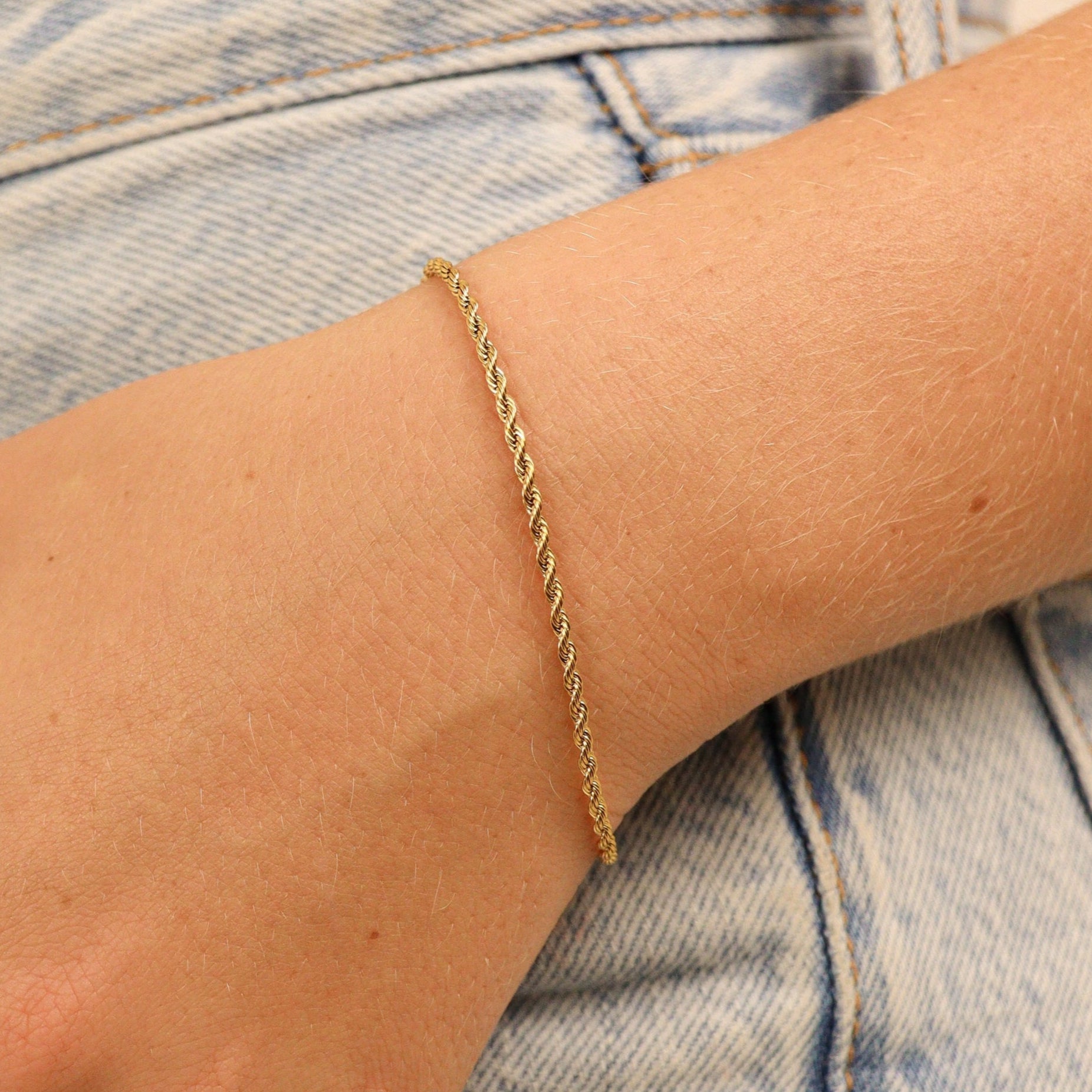 lose-up of a dainty 3mm gold rope chain bracelet styled on wrist with light wash denim for a casual look