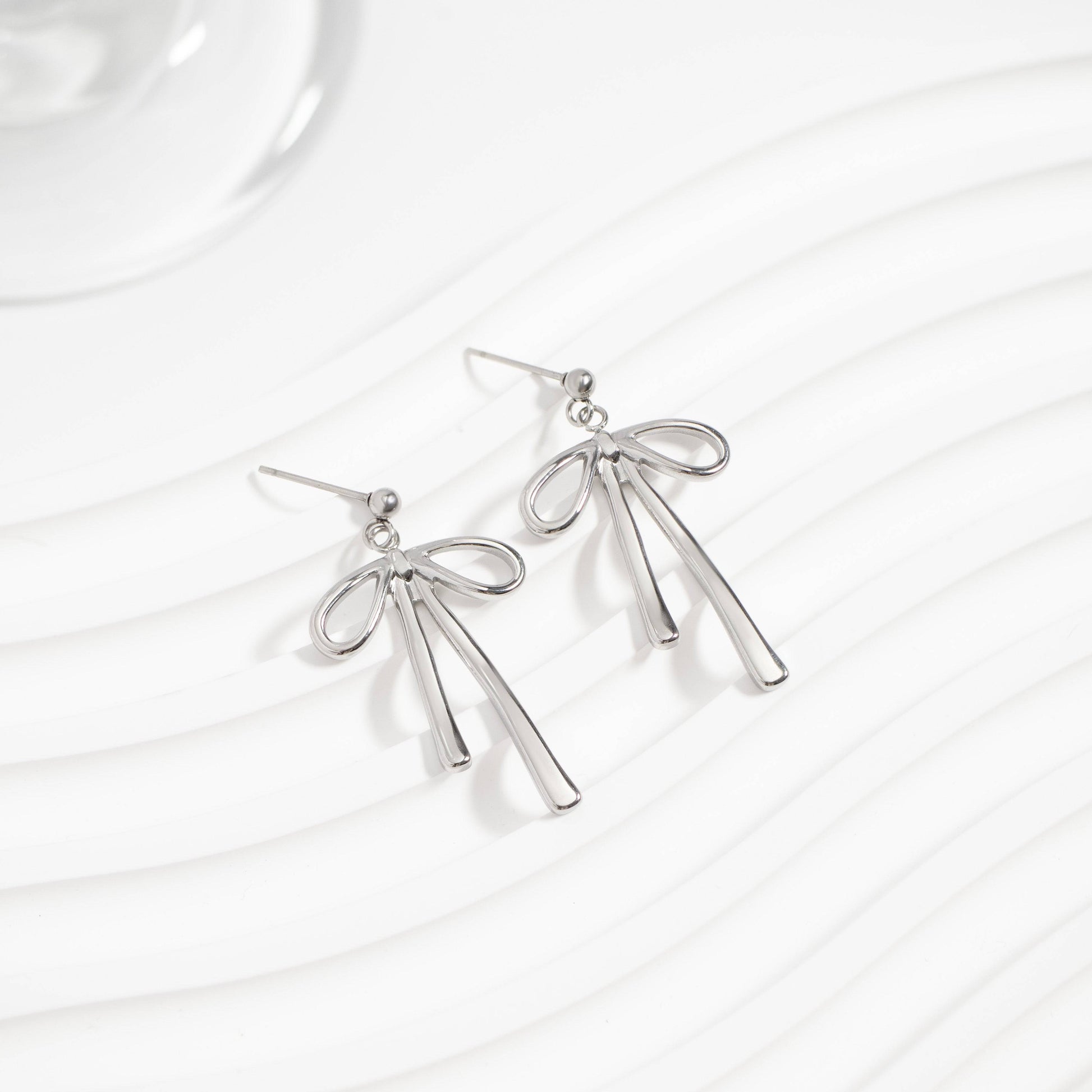 Pair of silver bow earrings displayed on a wavy white background. Dainty ribbon-inspired design that's hypoallergenic, tarnish-resistant, and perfect for everyday elegance from Lauren Riley Jewelry.