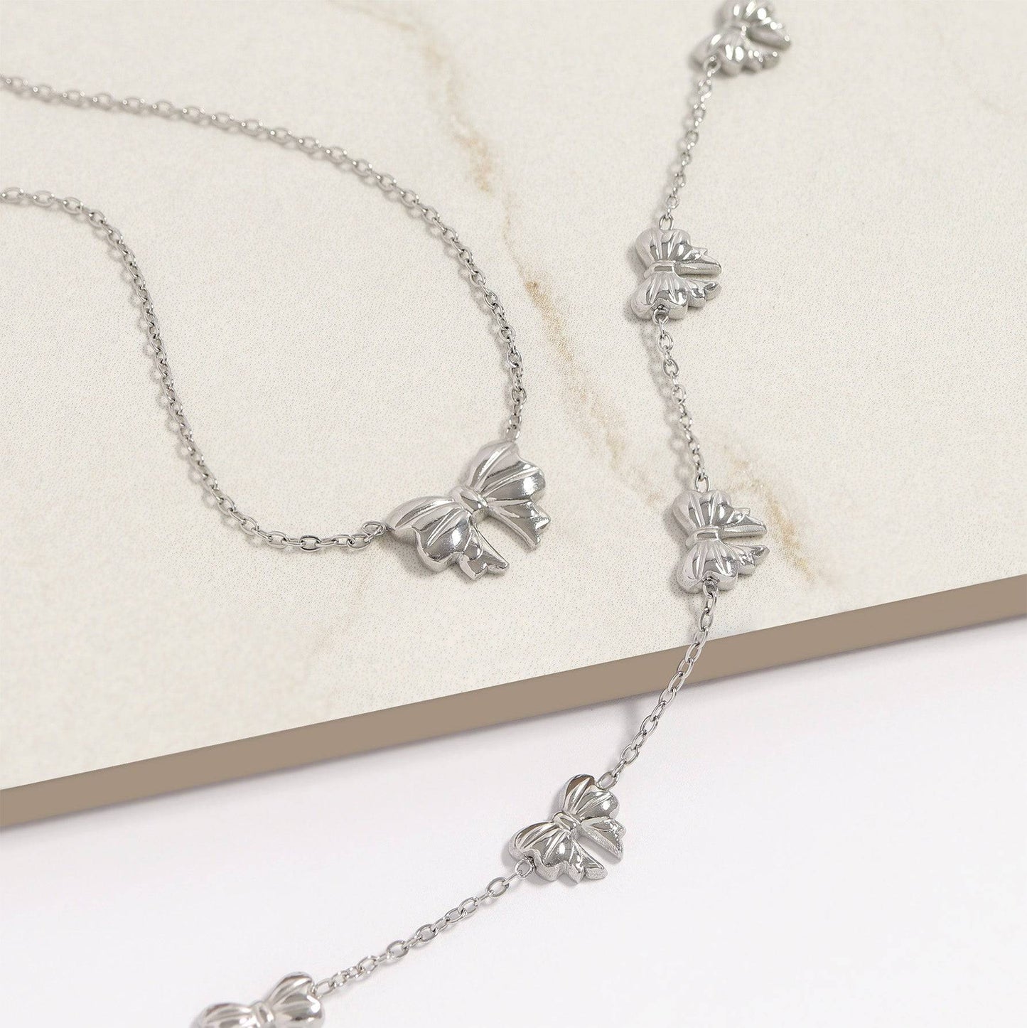 Silver bow necklace and bracelet set with dainty chains, waterproof and hypoallergenic stainless steel jewelry on neutral background.