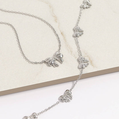Silver bow necklace and bracelet set with dainty chains, waterproof and hypoallergenic stainless steel jewelry on neutral background.