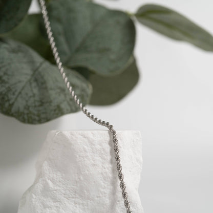 Stainless steel silver rope chain necklace draped over white rock with eucalyptus background, showcasing minimalist jewelry styling.